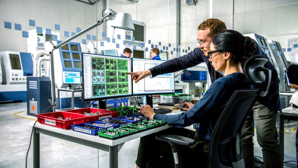 Engineers at a workstation reviewing PCB designs on monitors with organized component bins on the bench