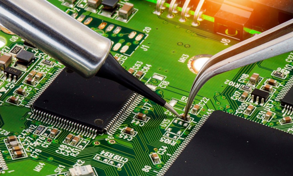Close-up of a soldering iron tip and precision tweezers reworking a fine-pitch surface-mount chip on a green PCB